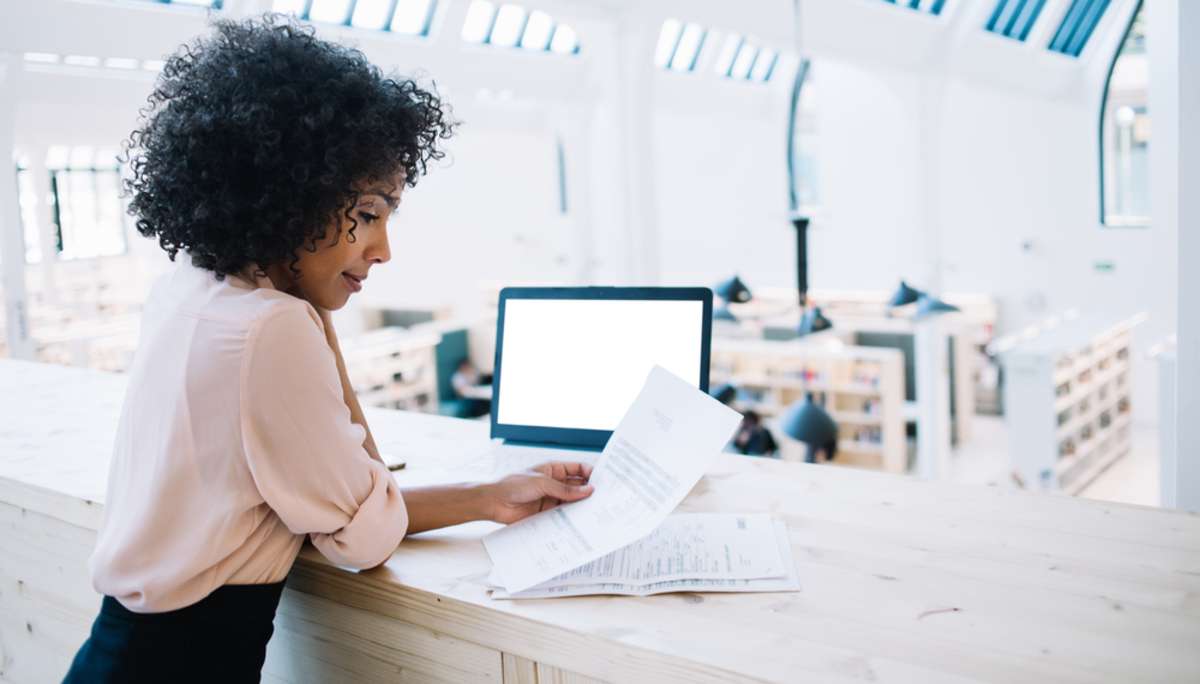 Side view of modern African American female reading paper documents while standing at counter with laptop